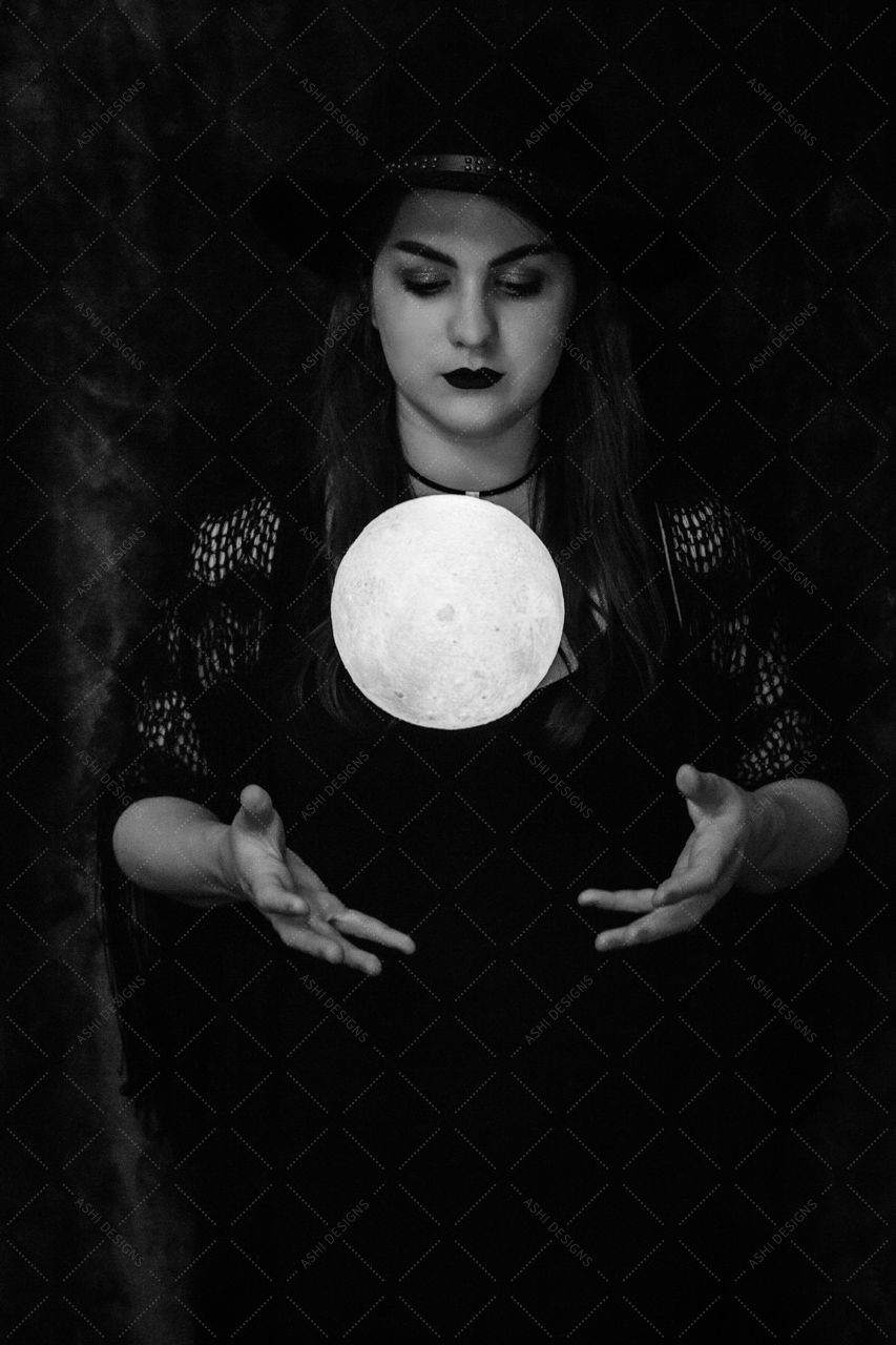 Black and white photo of Liz Loftus dressed in black, standing in a dark room, holding her hands out as a small white moon levitates above them.